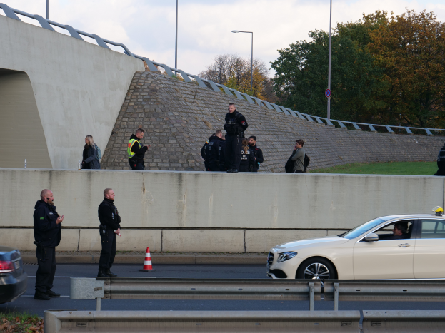 Eine Gruppe von Polizeibeamten steht neben einem Auto auf der Straße, mit Verkehrskegeln, einer Begrenzung, Gras, einer Wand, Laternen, Bäumen und einem bewölkten Himmel im Hintergrund.