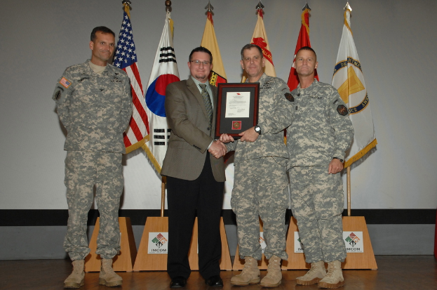 Vier Männer stehen auf einer Bühne: drei in Armeeuniformen und einer in Blazer, Krawatte und Hemd, die Hände schüttelnd und einen Preis überreichend, mit sechs Fahnen an Stangen und einer weißen Wand im Hintergrund.