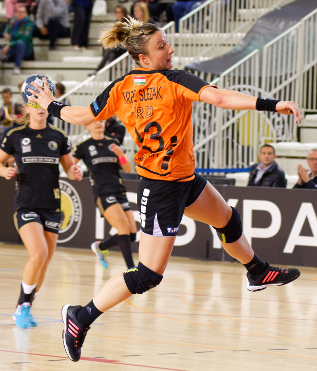 Eine Frau in einem orangen T-Shirt und schwarzen Shorts spielt Handball, hält einen Ball und hat Zuschauer und ein Banner mit der Aufschrift "Krp Slezak - Handball-Weltmeisterschaft 2019" im Hintergrund.