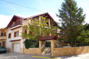 Eine Villa mit einem vor ihr geparkten Auto, das Fenster, Geländer, Säulen und ein Dach hat und von einem Tor, einem Schild, einem Straßenpfahl, Bäumen und einer Wand umgeben ist, unter einem bewölkten Himmel.