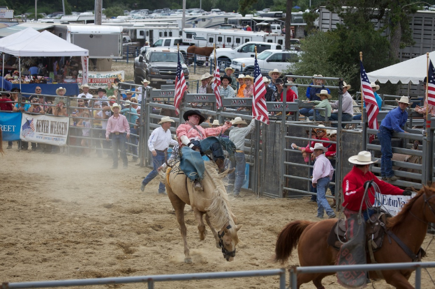 Zwei Reiter auf Pferden nehmen an einem Charreada-Event teil, umgeben von Zuschauern, hängenden Fahnen und Planen, parkenden Fahrzeugen und Bäumen im Hintergrund.