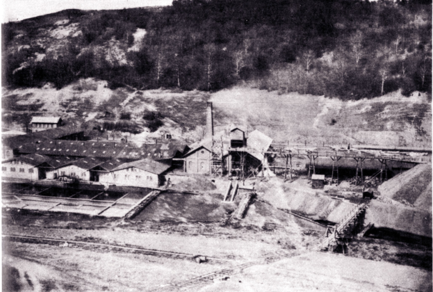 Schwarzes und wei├čes Foto eines Kohlebergwerks mit Geb├Ąuden, Masten und B├Ąumen im Hintergrund und Text unten.