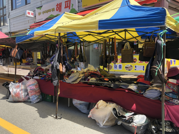 Ein Markt mit Zelten, die Taschen, Hüte und andere Gegenstände präsentieren, vor Häusern mit Fenstern und Texttafeln im Hintergrund.