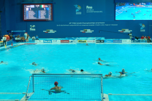 Eine Gruppe von Menschen spielt Wasserball in einem großen Pool mit einem Netz in der Mitte, Zuschauer auf der linken Seite und ein Display mit Text und Logos im Hintergrund bei den FINA-Weltmeisterschaften 2016 in Kazan.