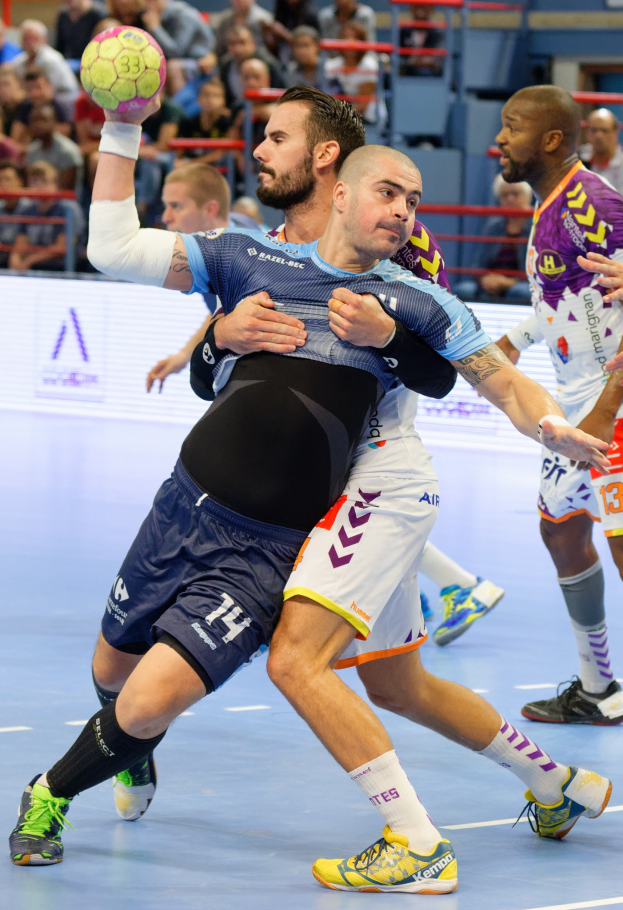 Eine Gruppe von Männern, die Handball in einem Stadion spielen, wobei einer den Ball hält und Zuschauer im Hintergrund sitzen.