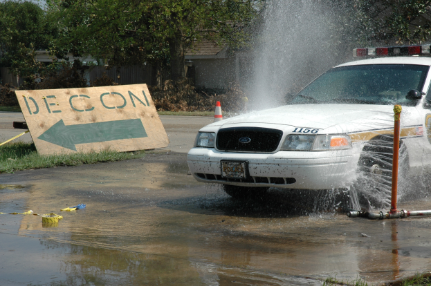 Ein Polizeiauto fährt durch eine Pfütze, mit einem Schild, einer Verkehrskegel und anderen Gegenständen auf dem Boden, vor einer Kulisse aus Bäumen, Gebäuden und einem klaren blauen Himmel.