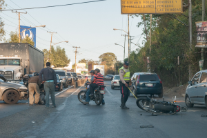 Eine Gruppe von Menschen steht um ein verunglücktes Motorrad auf der Straßenseite mit mehreren Fahrzeugen, darunter ein Lastwagen, und einer Hintergrund von Bäumen, Masten, Lichtern und Schildern unter dem Himmel.