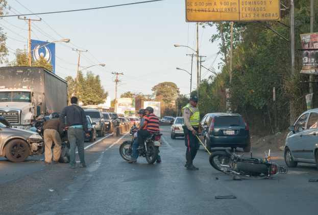 Eine Gruppe von Menschen steht um ein verunglücktes Motorrad auf der Straßenseite mit mehreren Fahrzeugen, darunter ein Lastwagen, und einer Hintergrund von Bäumen, Masten, Lichtern und Schildern unter dem Himmel.