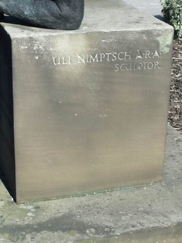Ein Stein mit Text in der Vordergrund, möglicherweise eine Statue oben, trockene Zweige, Erde und Pflanzen rechts, mit einer Straße im Hintergrund.