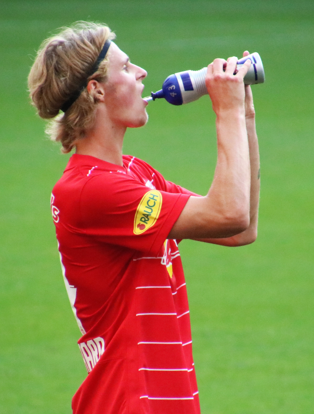 Ein Mann in rotem T-Shirt trinkt aus einer Flasche, während er auf einem Fußballfeld steht.