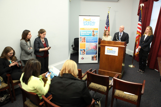 Drei sitzende Frauen, ein Mann und eine Frau vor einem Rednerpult, zwei Fahnen, eine Fahne, zwei Frauen links, eine Frau schaut auf eine Digitalkamera und Vorhänge oben rechts.