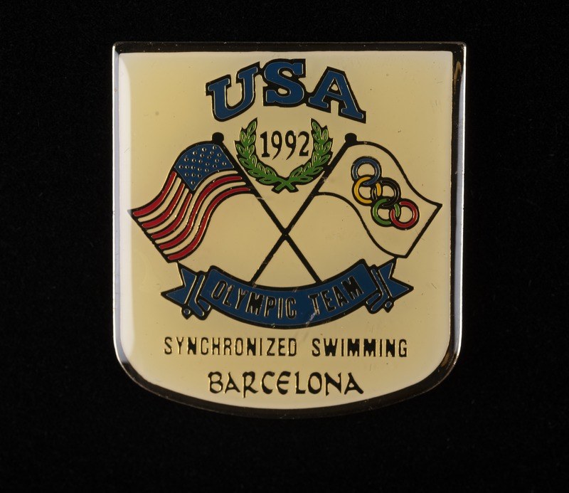 Abzeichen mit dem Text "USA Olympic Team Synchronized Swimming Barcelona" und zwei Flaggen (rot und blau) auf einem schwarzen Hintergrund.