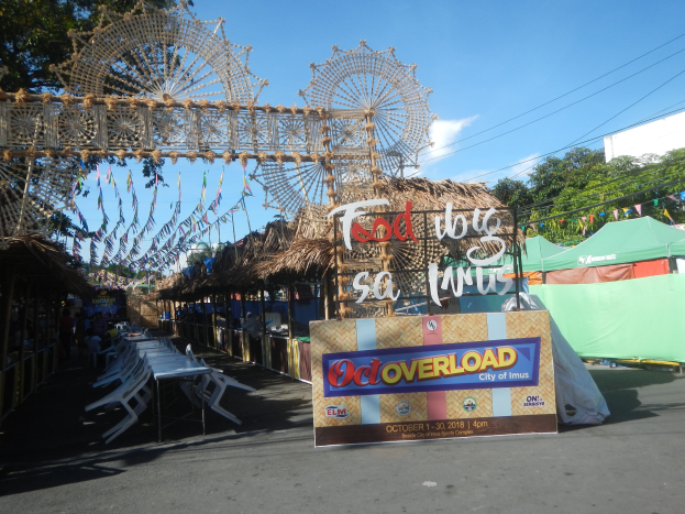 Eine KarnevalsSzene mit einem großen Riesenrad in der Mitte, umgeben von Hütten, Tischen, Stühlen, einem Schild, Dekorationen, Bäumen, Stromleitungen und einem bewölkten Himmel.