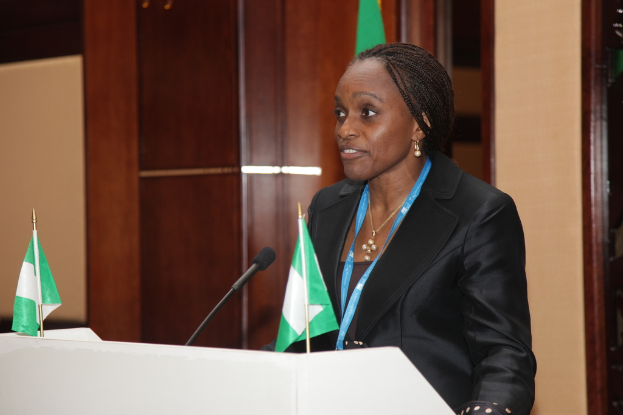 Eine Frau steht an einem Podium mit zwei Flaggen und einem Mikrofon, vor einer hölzernen Wand im Hintergrund.