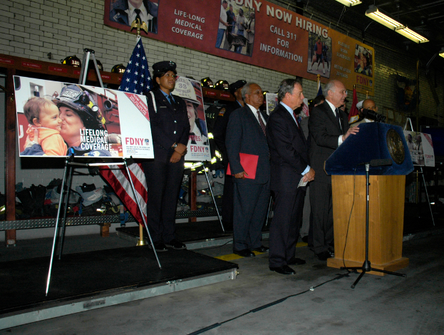 Eine Gruppe von Menschen steht in der Nähe eines Podiums mit Mikrofonen, Schildern auf Ständern, Fahnen und Schildern an der Wand, alles unter hellen Lichtern.