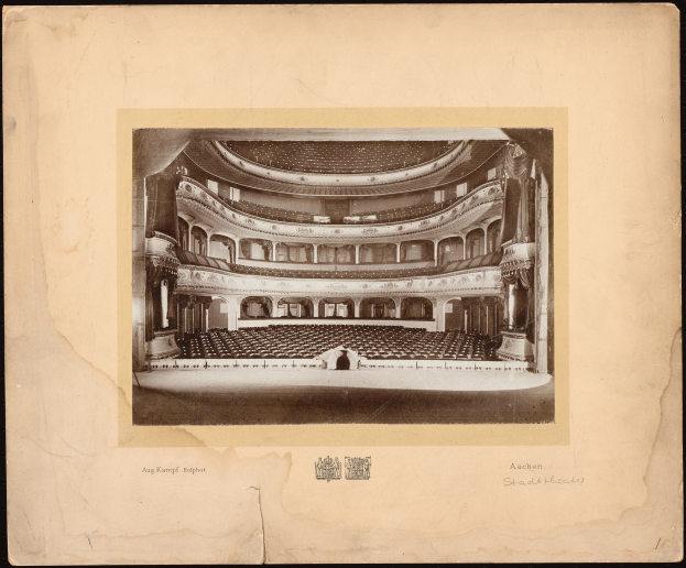 Altes Schwarz-Weiß-Foto des Auditoriums der Royal Albert Hall in London, das die Bühne, Sitzreihen, Säulen, Geländer und Text mit einem Logo unten zeigt.