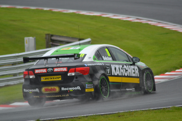 Ein Auto mit Text an der Seite, das auf einer regennassen Rennstrecke fährt, eingeräumt von Gras, mit einem Geländer und einer hölzernen Struktur im Hintergrund.