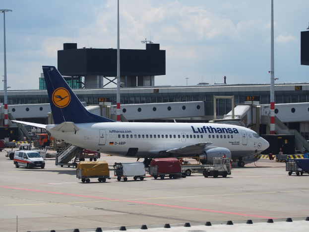 Lufthansa Airbus A320-200 am Frankfurt Airport mit Bodenfahrzeugen und Wagen vorne, einem Geb"ude und Laternen im Hintergrund unter einem klaren blauen Himmel.
