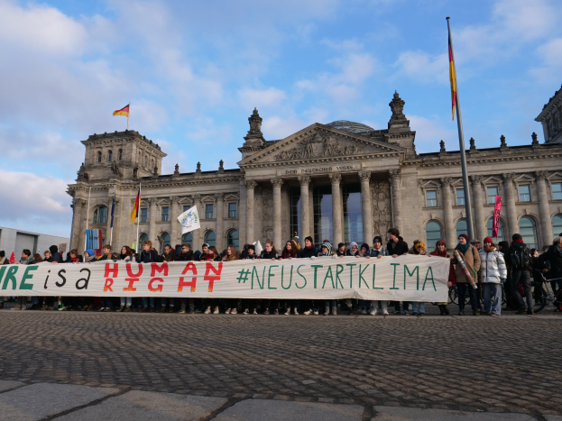 Gruppe von Menschen mit einem Banner 'Wir sind ein Menschenrecht' vor dem Reichstaggebäude, das Säulen, Fenster, Bögen und Statuen sowie Flaggen und einen bewölkten Himmel im Hintergrund zeigt.