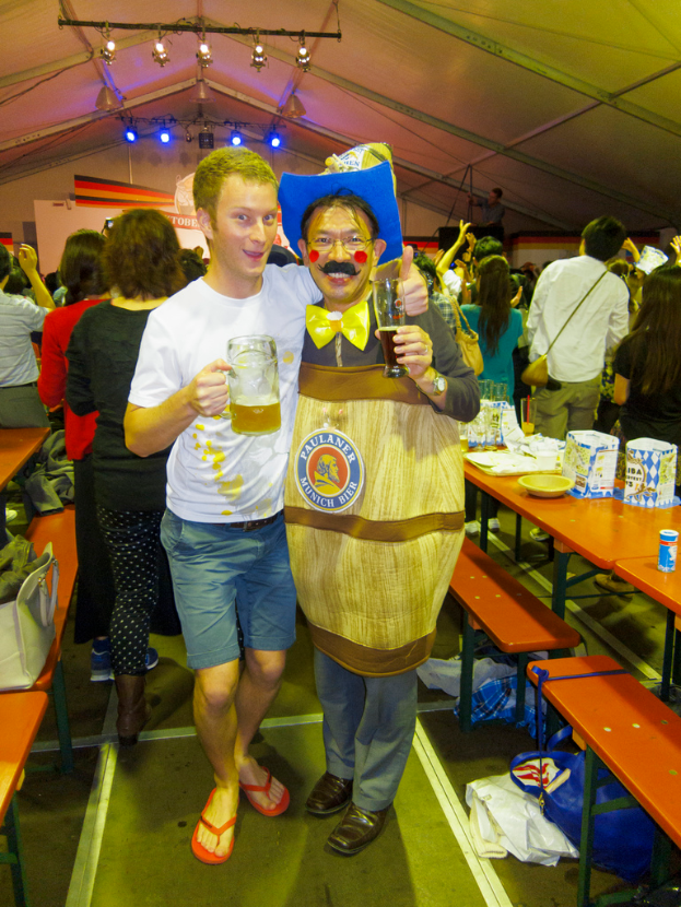 Zwei Personen stehen zusammen, eine hält ein Kind mit einem Bier, die andere in einem Dramakostüm, mit einem Tisch voller Essen daneben und einer Gruppe von Menschen dahinter.