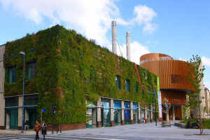 Ein großes Gebäude mit einer grünen Wand an der Seite, umgeben von Straßenmasten, Straßenlaternen, Verkehrspfosten, Verkehrssignalen, Kraftfahrzeugen, Fußgängern, Bäumen und einem bewölkten Himmel, als Teil des Green Building Project.
