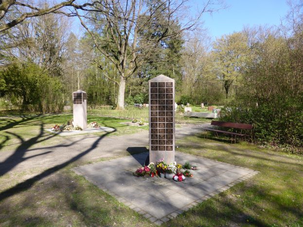 Kriegsdenkmal auf einer grünen Wiese mit Bäumen, umgeben von Blumensträußen und einer Bank, Gräber im Hintergrund unter einem klaren blauen Himmel.