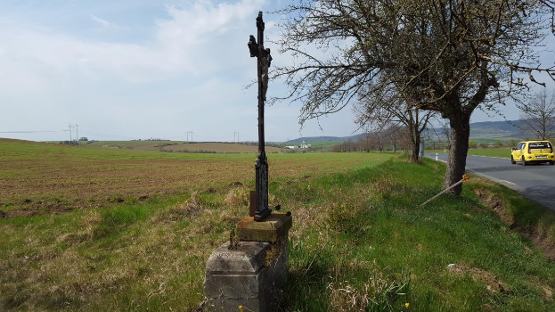 Ein gelbes Auto fährt auf einer Straße neben einem Baum, mit einem Kreuz am Straßenrand, Hügeln und einem bewölkten Himmel im Hintergrund und Gras und Blumen auf dem Boden.