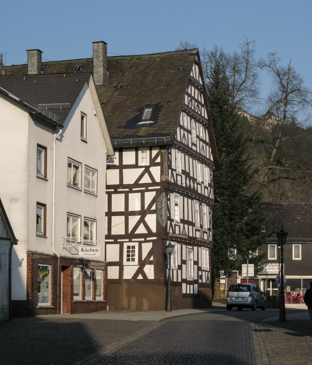 Holzhaus in einer Kleinstadt umgeben von Gebäuden, Fahrzeugen, Fußgängern, Bäumen und Straßeninfrastruktur unter einem klaren blauen Himmel.