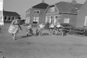 Eine Gruppe von Kindern, die auf Fahrrädern eine Straße entlangfahren, mit einer Frau, die neben ihnen geht, Straße mit Pflanzen und einem Zaun gesäumt, Gebäude mit Fenstern im Hintergrund unter einem klaren Himmel.
