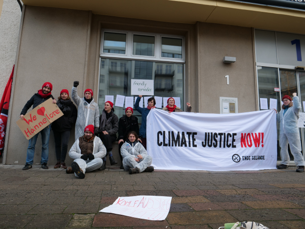 Eine Gruppe von Menschen mit Mützen hält ein Transparent mit der Aufschrift "Climate Justice Now" vor einem gläserwändigen Gebäude, mit Papieren, einer Flagge und einer Tasche auf dem Boden.