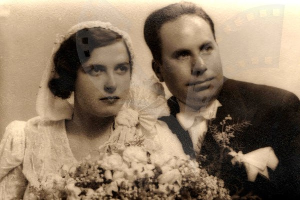 Ein Schwarz-Weiß-Foto von einem Mann und einer Frau, die zusammenstehen, die Frau in einem weißen Kleid mit einem Strauß Blumen, beide lächeln.