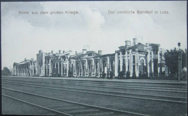 Ein altes Schwarz-Weiß-Foto eines Bahnhofs in Lida, Deutschland, das ein großes Gebäude mit Säulen, eine Gruppe von Menschen, Pfosten, einen Zaun, einen Straßenpfosten, Bäume und einen bewölkten Himmel zeigt.
