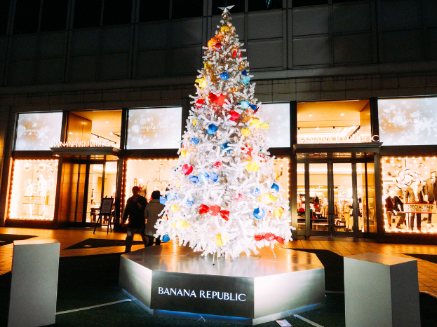 Ein Baum mit Geschenken darauf auf einer Plattform, mit einem Gebäude, Türen, Lichtern, Kleidern und anderen Gegenständen im Hintergrund und zwei Personen auf der Straße.