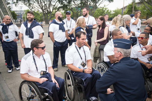 Eine Gruppe von Menschen in Rollstühlen unterhält sich vor einem Hintergrund aus Bäumen, Pfählen, Gebäuden und einem klaren blauen Himmel, wobei einige Mützen und Brillen tragen, was darauf hindeutet, dass sie bei den Paralympischen Spielen sind.