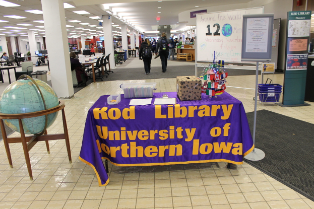 Eine Expo-Situation mit einem Tisch, auf dem verschiedene Landesflaggen, ein Karton, Papiere und ein Banner liegen, eine Kugel links daneben, ein Anschlagbrett rechts mit ein paar Menschen in der Nähe und mehreren Tischen und Stühlen ganz links.