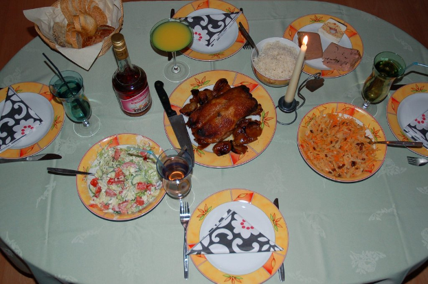 Ein Tisch mit Tellern, Gläsern, einer Flasche, Schüssel, Korb, Messern, Löffeln, einer Kerze und verschiedenen Lebensmitteln.