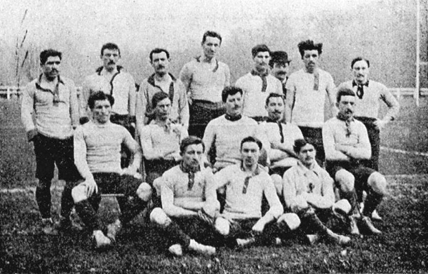 Ein Schwarz-Weiß-Foto einer Fußballmannschaft aus den 1920er Jahren, mit einigen Spielern, die stehen, und anderen, die auf dem Boden sitzen, vor einer Kulisse aus Pfählen, Bäumen und einem klaren Himmel.