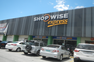 Ein Shopwise Express-Laden mit mehreren Autos davor, der eine Fahne auf der linken Seite trägt, unter einem bewölkten Himmel.