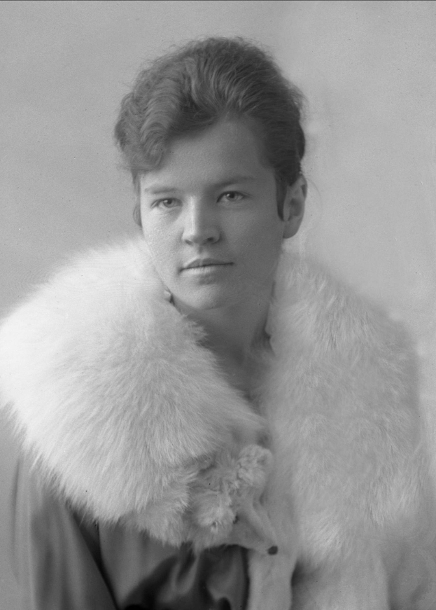 Ein Schwarz-Weiß-Foto einer Frau mit ernstem Gesichtsausdruck, deren Haar zurückgebunden ist und die einen Pelzmantel trägt, der um ihre Schultern liegt und in die Ferne schaut.