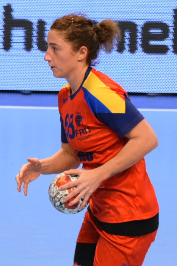 Eine Frau in einer orange-blauen Uniform hält einen Fußball vor einem Bildschirm mit Text.