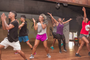 Eine Gruppe von Menschen tanzt in einem Tanzstudio mit einem Gemälde an der Wand und einem Glasfenster auf der rechten Seite, unter Deckenbeleuchtung, während einer Zumba-Klasse.