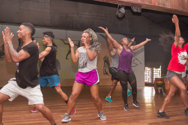 Eine Gruppe von Menschen tanzt in einem Tanzstudio mit einem Gemälde an der Wand und einem Glasfenster auf der rechten Seite, unter Deckenbeleuchtung, während einer Zumba-Klasse.