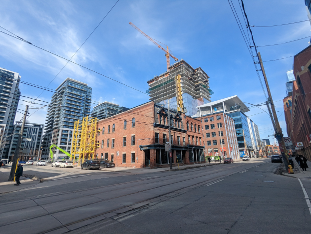 Eine belebte Stadtstraße in Toronto mit fahrenden Fahrzeugen, Fußgängern auf dem Gehweg, Oberleitungen, Schildern, Gebäuden und einem Baustellenschild im Hintergrund bei bewölktem Himmel.