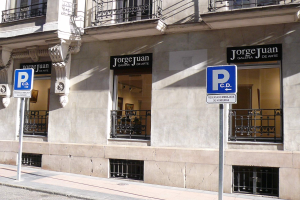 Ein mehrstöckiges Gebäude mit Fenstern, Geländern, Türen und Säulen, das Schilder an Pfosten entlang des Gehwegs und einsehbare Innenobjekte zeigt, die eine belebte Stadtkulisse suggerieren.