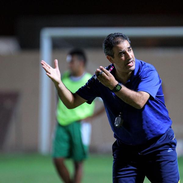 Ein Mann in einem blauen T-Shirt und Hose steht auf einem Fußballfeld und hält ein Mikrofon in der Hand, während er als Trainer gestikuliert. Im Hintergrund sind ein Torpfosten, eine Wand und eine weitere Person in einem grünen Kleid zu sehen.
