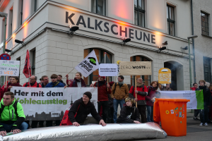 Eine Gruppe von Menschen steht vor einem Gebäude und hält Schilder und Plakate in die Höhe, während zwei Personen im Vordergrund sitzen und ein Müllcontainer rechts zu sehen ist, während sie in Deutschland protestieren.