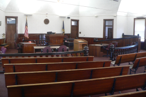 Ein Gerichtssaal mit hölzernen Kirchenbänken, Tischen, Stühlen, einem Podium, Fahnen, Türen, einer Uhr an der Wand und Lampen und Ventilatoren an der Decke.