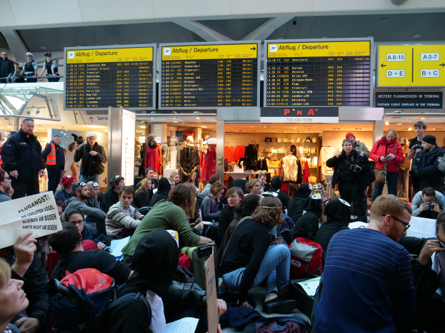 Eine große Gruppe von Menschen in einem Flughafen, einige sitzen mit Taschen und Papieren, während andere stehen, mit Texttafeln, Schaufensterpuppen in Kleidern und Deckenleuchten im Hintergrund, was auf eine Demonstration hinweist.