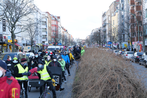 Eine große Gruppe von Menschen in Masken und Sicherheitswesten auf Fahrrädern auf einer von Bäumen gesäumten Straße mit Gebäuden, Lichtmasten und Texttafeln, neben Fahrzeugen und trockenem Gras, unter einem klaren blauen Himmel.