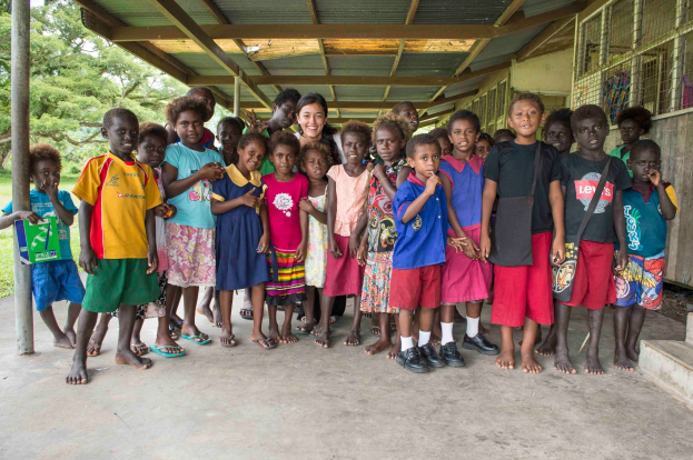 Eine Gruppe von Kindern steht vor einem Gebäude, mit einer Frau in der Mitte, Treppe auf der rechten Seite, ein Pfahl auf der linken Seite und üppiger Vegetation im Hintergrund.